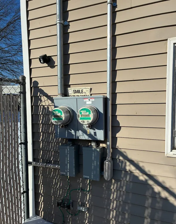 Two utility meters in a gray double meter socket on tan lap siding, fed by galvanized service risers with disconnect boxes below