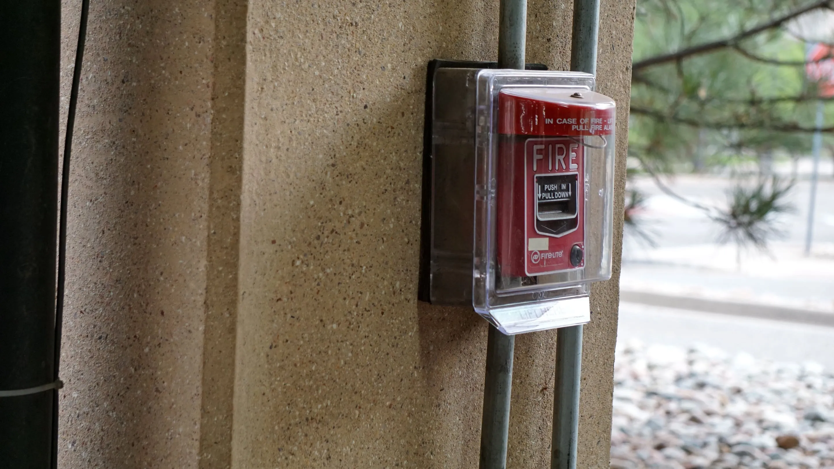 Red fire alarm pull station under a clear protective cover, fed by metal conduit on a concrete wall